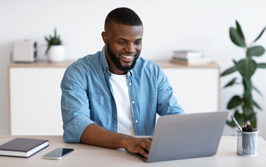 Wall Mural - Distance Job. Millennial African American Man Working Online On Computer At Home Office, Handsome Smiling Black Freelancer Guy Sitting At Desk And Looking At Laptop Screen, Free Space