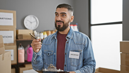 A bearded man volunteers at an indoor workplace, holding a clipboard in a denim jacket labeled 'volunteer'.