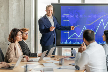 Aufkleber - A businessman stands in front of a large screen displaying a graph with an upward trend, pointing to the data. He is presenting to a group of colleagues seated at a conference table.