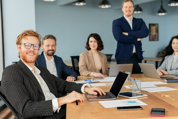 Aufkleber - A team of business professionals is gathered around a large conference table, collaborating and working on projects. One man, with a red beard and glasses, is smiling and working on a laptop