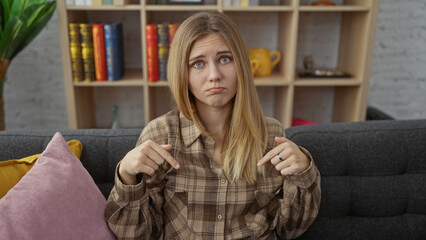 Wall Mural - Confused young woman with blonde hair sitting on a couch indoors, pointing at herself in a living room.