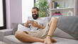 © Krakenimages.com - Handsome bearded hispanic man relaxing and smiling while using his smartphone on a comfy sofa in a modern living room.