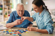 © pikshine - A nurse is helping an elderly man do puzzle games at home, they smile and have fun together. Old age, dementia, Alzheimer's disease, therapy concept