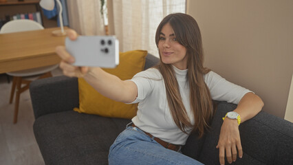 Sticker - A smiling young hispanic woman takes a selfie in a well-lit living room, conveying a casual and comfortable atmosphere.