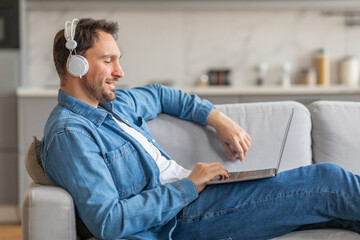Poster - A man in a denim shirt sits on a light-colored couch, relaxed with headphones on and his laptop in his lap. He is smiling, looking at the screen of his computer. The room has a modern design