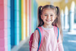 © AISTEL - cheerful little brunette girl student with backpack walking through the elementary school hallway, confidently smiling at the camera. Back to school concept. copy space for text.