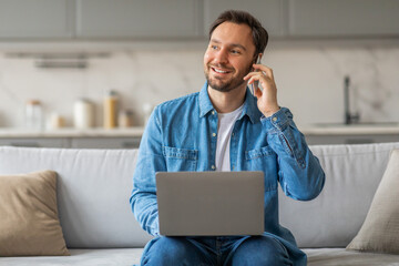 Poster - A man wearing a blue shirt and jeans sits on a white couch in a living room. He is smiling and talking on his phone with a laptop in his lap. There is a throw pillow on the couch beside him.
