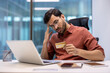 © Liubomir - Man in office experiencing stress holding credit card while looking at laptop. Concept of financial issues, online transaction error, office work. Professional setting with modern workspace