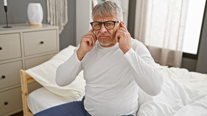 Sticker - Mature grey-haired man covering his ears while sitting on a bed indoors, showing discomfort or needing quiet.