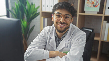 Wall Mural - Handsome young arab man with beard in white lab coat smiling in indoor clinic office setting