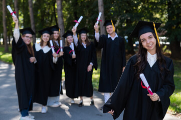 Wall Mural - Group of happy students in graduation gowns outdoors. A young girl with a diploma in her hands in the foreground.