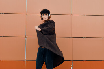 Wall Mural - Fashionable young man in headphones, wearing a brown coat and black pants, posing against an orange wall with his arms out to the side