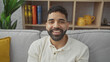 © Krakenimages.com - A smiling young hispanic man with a beard sitting on a couch in a cozy living room with bookshelves