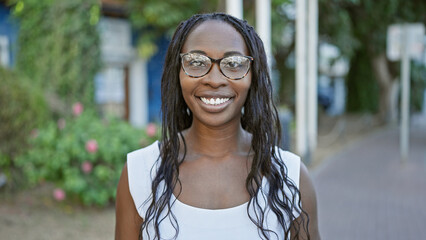 Wall Mural - Smiling young adult african american woman with curly hair and glasses posing outdoors in a park.