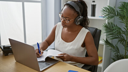 Wall Mural - African american woman wearing headphones focuses on taking notes during her work in a modern indoor office.