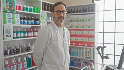 Poster - A mature hispanic man in a white coat stands confidently inside a pharmacy full of medical supplies.