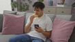 © Krakenimages.com - A handsome young hispanic man in casual clothing eats a snack while using a smartphone on the couch in a modern living room setting.
