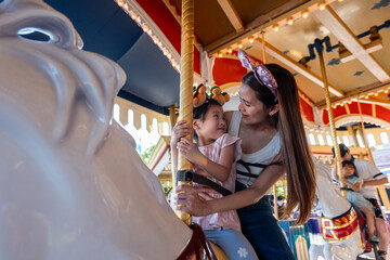 Asian mother and adorable kid daughter riding carousel at theme park.