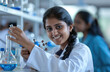 © Kien - A smiling young Indian female scientist is holding and looking at a model of a molecule with red balls on one end, blue liquid in beakers behind her and test tubes around it.