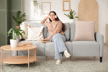 Wall Mural - Beautiful young woman in headphones listening to audiobook with books and sitting on sofa in living room