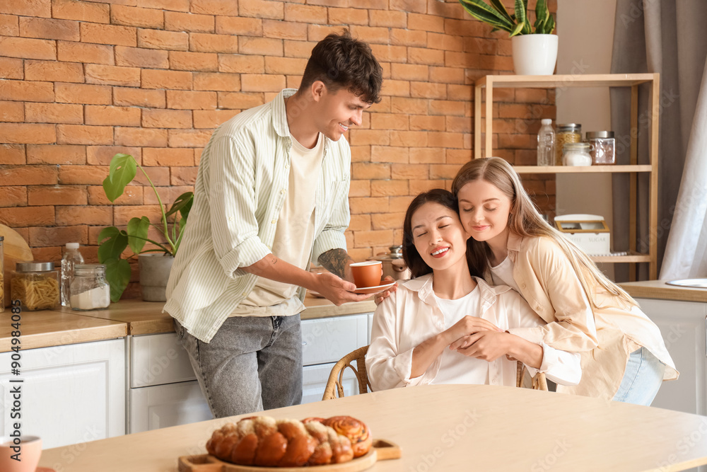 Young man with coffee cup and two women hugging in kitchen. Polyamory concept