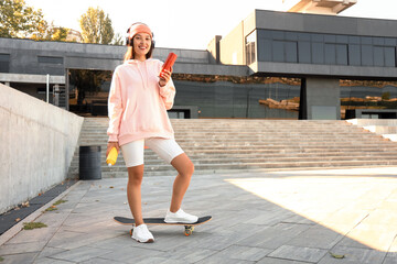 Wall Mural - Young Asian woman in headphones with spray paint cans skating on street