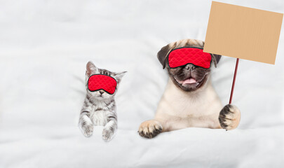 Fototapete - Pug puppy and tiny kitten wearing sleeping masks sleep together on a bed at home. Pug puppy  shows blank placard. Top down view. Empty space for text