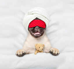 Fototapete - Pug puppy wearing sleeping mask sleeps with toy bear under white blanket on a bed at home. Top down view