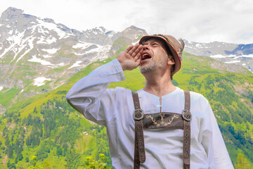 A man in traditional Tyrolean national dress shouts on a mountain ridge in the Alps in daylight