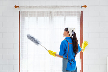 Asian young professional cleaning service woman worker working in the house. The girl cleans the curtain and window with a grey feather duster. Professional cleaner and chore service concept.