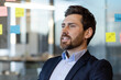 © Liubomir - Confident business professional in modern office setting with colorful sticky notes on glass wall. Man in blazer looking forward with positive expression, representing leadership and strategy.