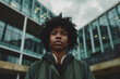 © yaqui_villegas - A young individual with curly hair and headphones around their neck stands confidently outside a modern building, ready to face the world with determination and style.