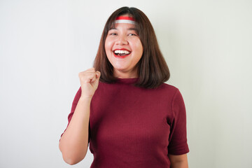 Excited young asian woman wearing Indonesian flag headband, smiling expression, hands raised with clenched fist gesture, isolated over white background. Concept for Indonesian Independence Day.