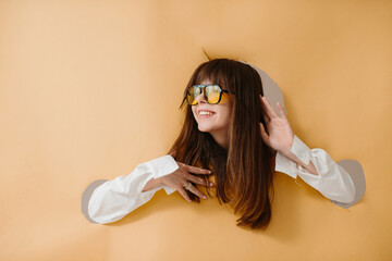 A sincere smiling young woman in sunglasses posing on a torn beige background.