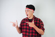 © Rizky - Excited amazed young asian man wearing Indonesian flag headband and pointing fingers on copy space beside, isolated over white background. Concept for Indonesian Independence Day.