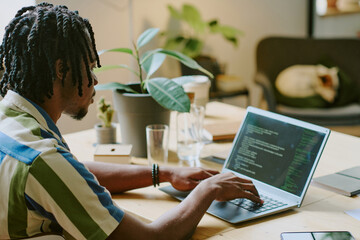 Young African American programmer wearing casual clothes coding on laptop in modern eco-friendly office, his dog sleeping on chair in background