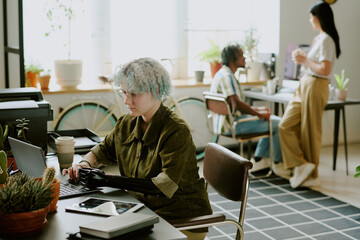 gen z caucasian girl with disability wearing arm prosthesis working on laptop, her colleagues having