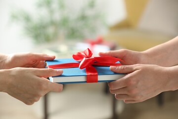 Wall Mural - Woman gifting her friend book tied with red ribbon indoors, closeup