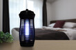 © Sutthisak - an insects mosquito electric blue light killer lamp is put on nice wooden table in nice bedroom with background of white bed and towel and digital clock to protect the mosquito during sleeping time
