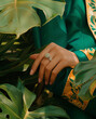 © sunaiart - A close-up of a hand with a diamond ring is set against the backdrop of an elegant green suit with yellow details, amidst a tropical setting in a jewelry photograph.