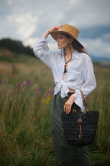 Wall Mural - Fashionable confident woman wearing trendy summer outfit with wide brim hat, white linen shirt, green skirt, holding black wicker  straw bag, posing outdoor.  Fashion portrait