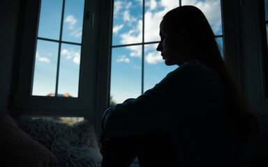 Close up side view of shadowed silhouette depressed woman while she sitting on windowsill with wide window on background. Sad, crisis, bad emotions, mental health, family violence.