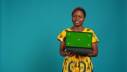 Wall Mural - Confident smiling woman presenting a chroma key display on laptop, standing against blue background. Young adult holding a wireless pc and showing a green screen mockup. Camera B.