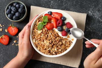 Wall Mural - Woman eating tasty granola with berries and yogurt at dark textured table, top view