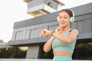 Poster - Sporty Asian woman in headphones with smartwatch checking pulse outdoors