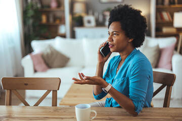 Smiling middle aged woman sitting on chair at home talking over smartphone. Happy black woman using mobile phone for conversation. 