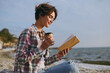 © ViDi Studio - Full body side view young woman she wears shirt casual clothes reading paper book novel drink coffee sits rest on sea ocean sand shore beach outdoor seaside in summer day free time. Lifestyle concept.