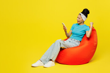 Wall Mural - Full body young Latin woman wears blue top t-shirt headband casual clothes sit in bag chair hold in hand use mobile cell phone do winner gesture isolated on plain yellow background. Lifestyle concept.