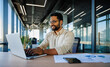 © Pixelowy_czarodziej - A smiling young man in a suit sits in the office at a desk and works on a laptop, typing on a keyboard, texting, working with data