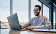 © Pixelowy_czarodziej - A smiling young man in a suit sits in the office at a desk and works on a laptop, typing on a keyboard, texting, working with data